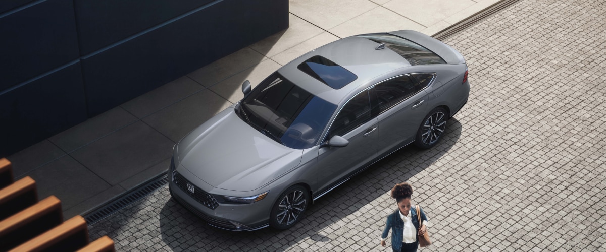 3/4 bird’s eye view of woman walking away from a grey Accord parked curbside in an upscale urban setting.