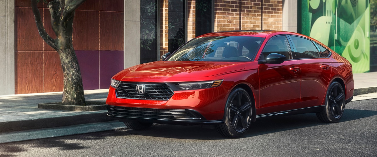 3/4 front view of red Accord parked curbside next to a tree outside of a trendy brick building with art on the window.