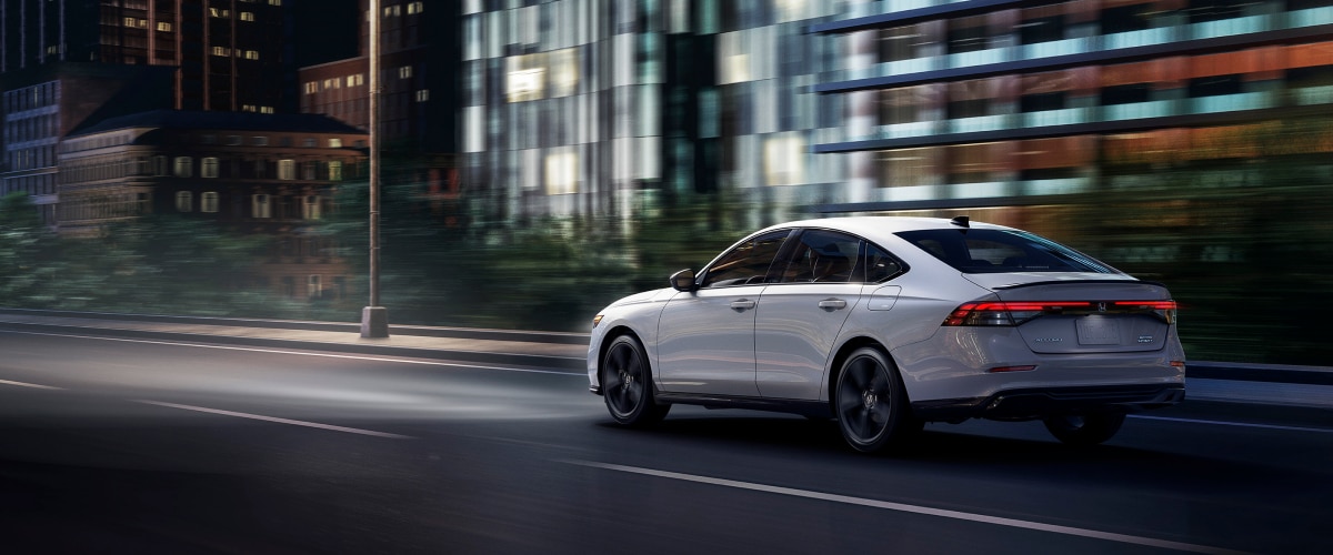 3/4 rear view of white Accord driving on city side highway past buildings at night.