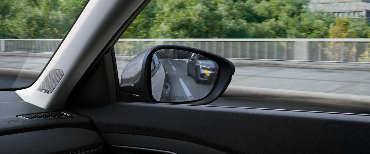 Closeup view of Interior view of door mirror with the Blind Spot Indicator light on, reflecting a car in the other lane.