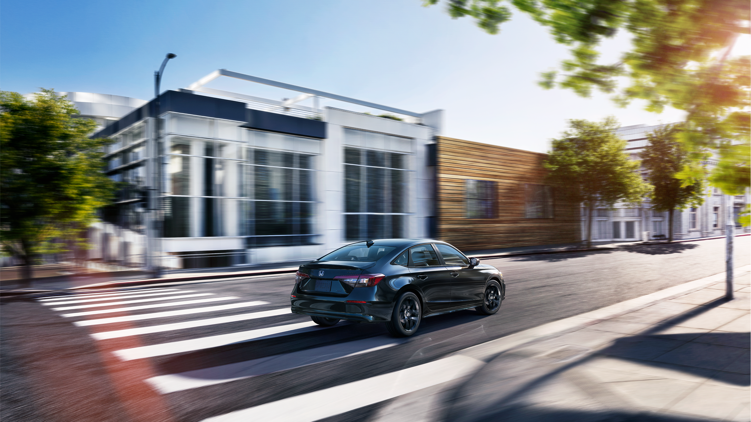 3/4 rear wide view of a black Civic Sedan driving by upscale boutique architecture on a city street during the day.