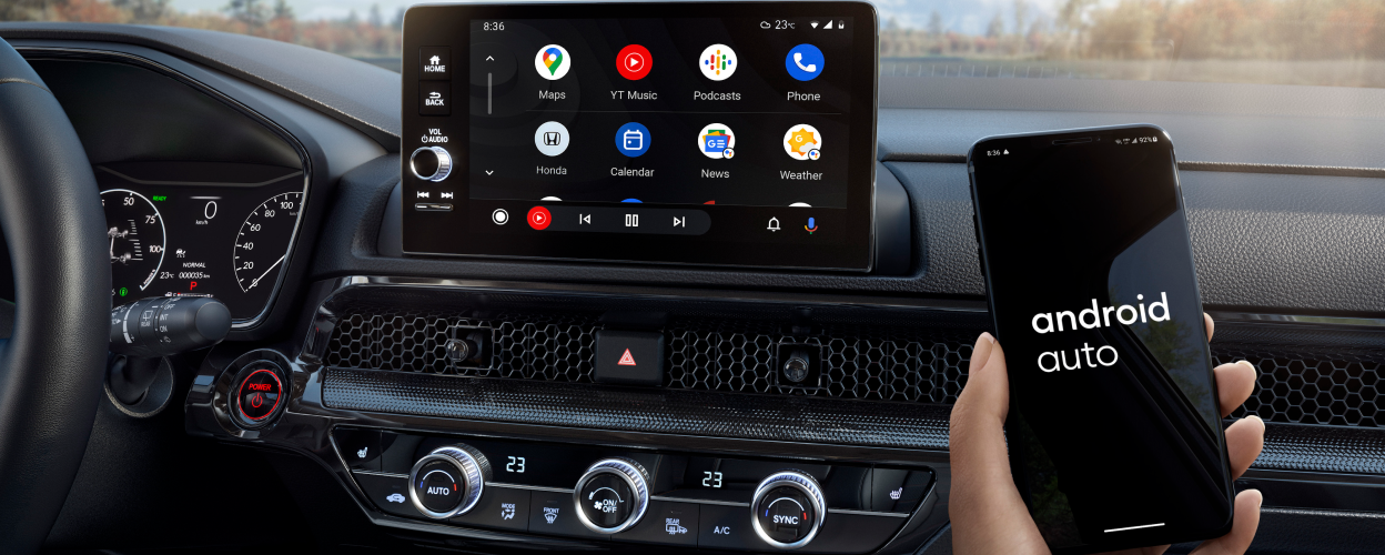 Gros plan de l’écran tactile. Au premier plan, une main tenant un téléphone intelligent sur lequel on peut lire « Android Auto » à l’écran. Forêt en arrière-plan dans le pare-brise. 