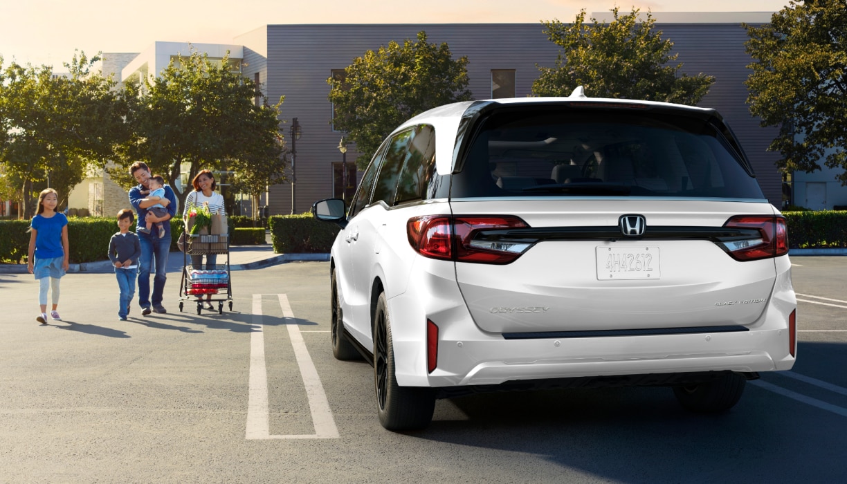 Rear view of grey Odyssey in empty parking lot. Family of walks toward it, pushing a grocery cart.