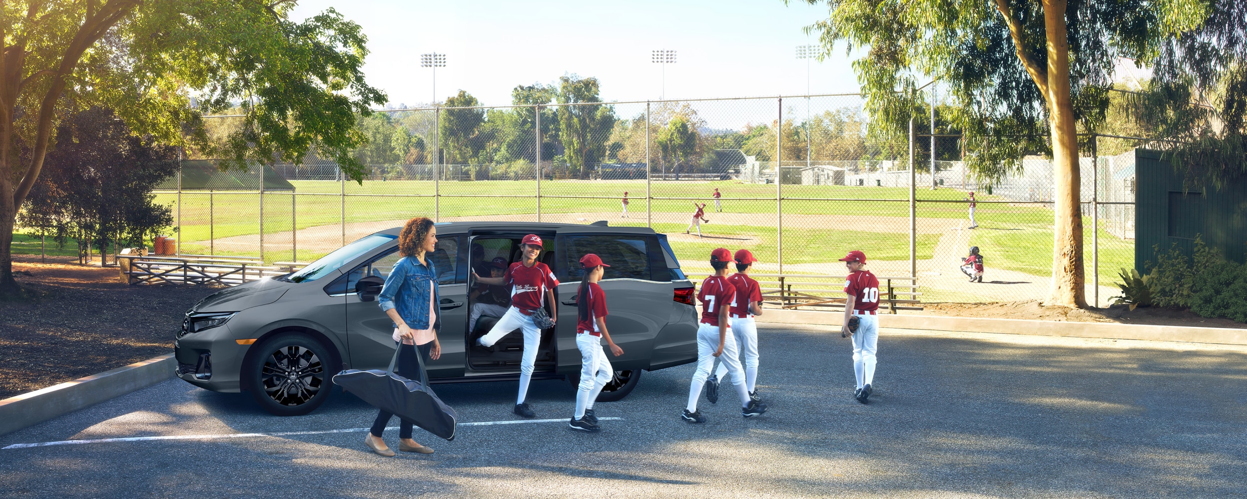 Mom and kids’ baseball team exiting a grey Odyssey parked near a baseball diamond.