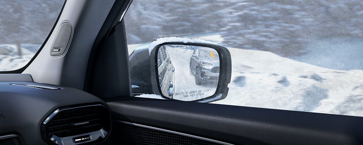 View of door mirror, seen from inside, showing a vehicle in the reflection and an illuminated alert light.