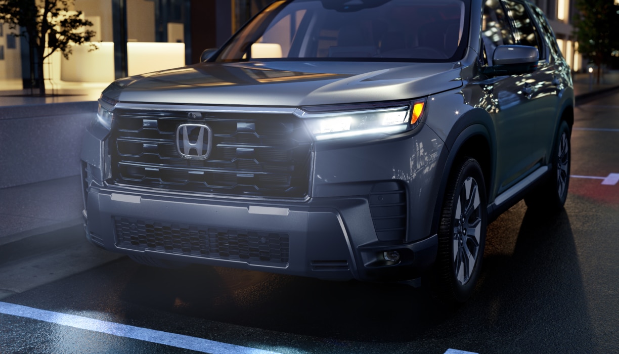Front view of a silver Pilot driving on a dark, snow-covered road with headlights on. Blowing snow surrounds the vehicle and a cargo box is mounted on the roof.