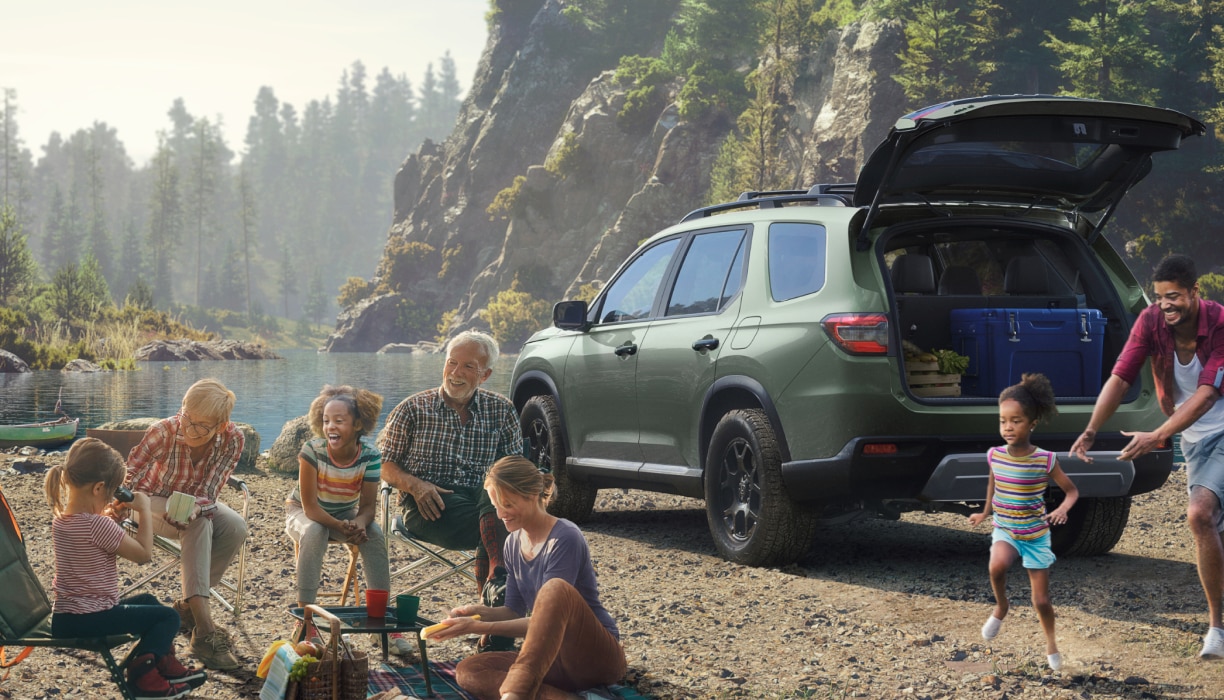 3/4 rear view of a green Pilot parked near a lakeside mountain. The tailgate is open, revealing a blue box and wooden crate inside. Nearby, a parent chases a young girl, while a family sits on folding chairs around a picnic blanket.