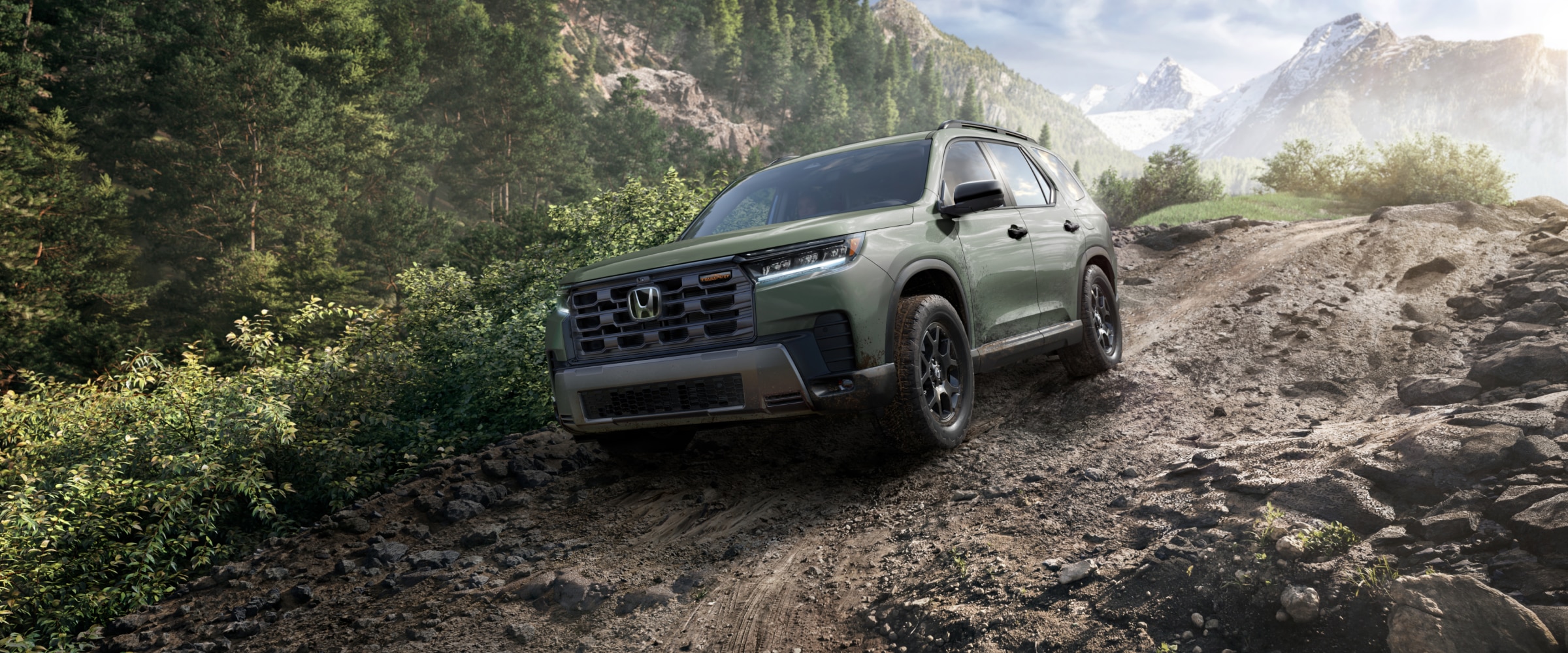 3/4 front side view of a green Pilot driving downhill on a dirt path in the forest, with distant mountains in the background.