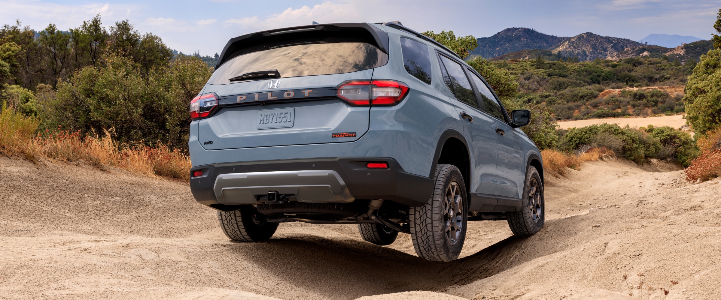 3/4 rear view of a light blue Pilot driving through uneven terrain with mountains in the background.