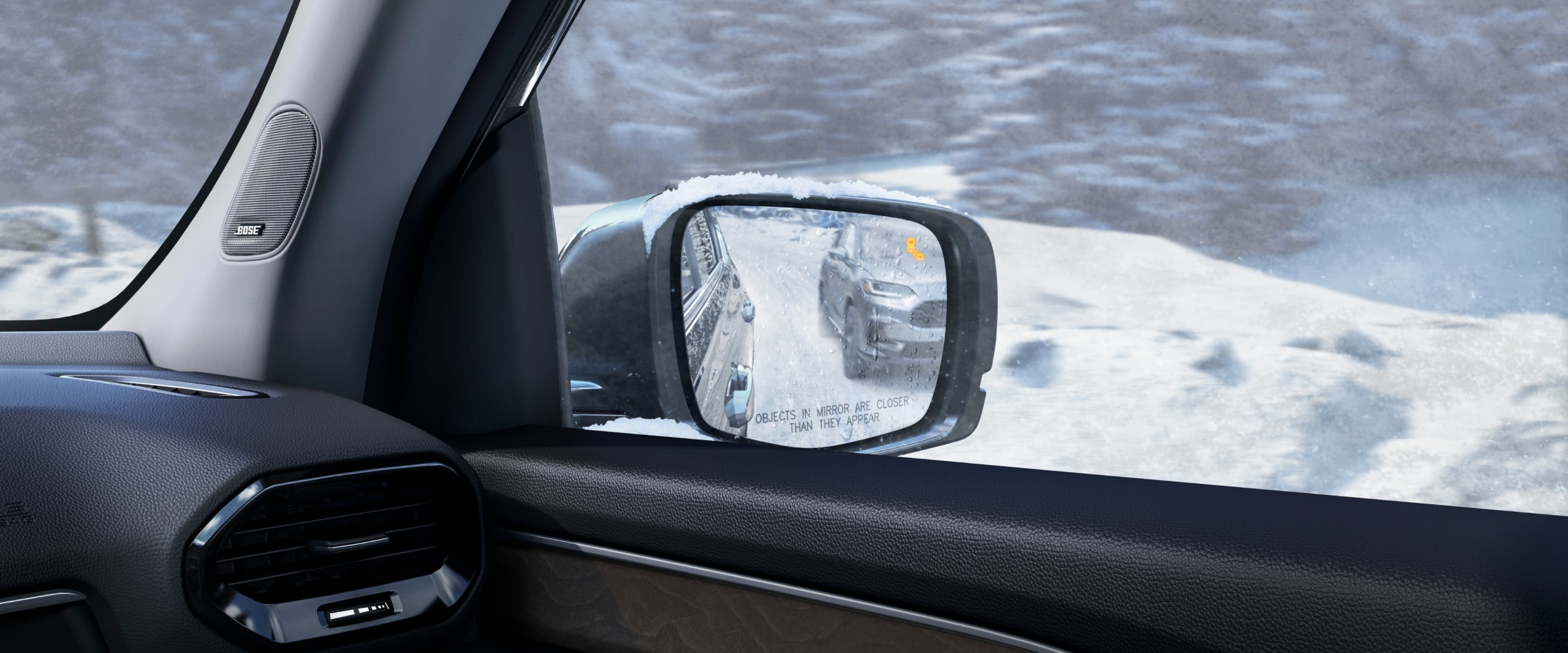 Passenger-side view of a snow-covered side mirror with an illuminated alert, reflecting a vehicle in the adjacent lane.