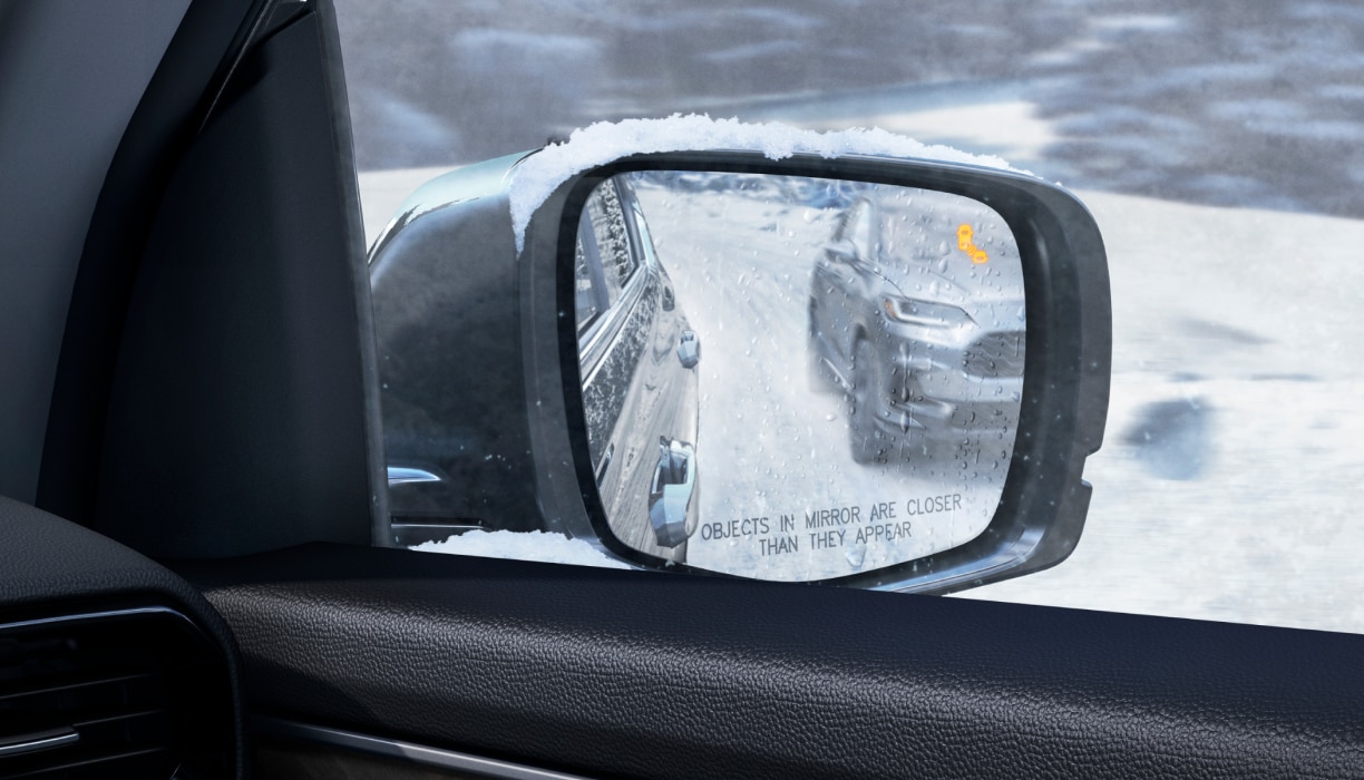 Passenger-side view of a snow-covered side mirror with an illuminated alert, reflecting a vehicle in the adjacent lane.