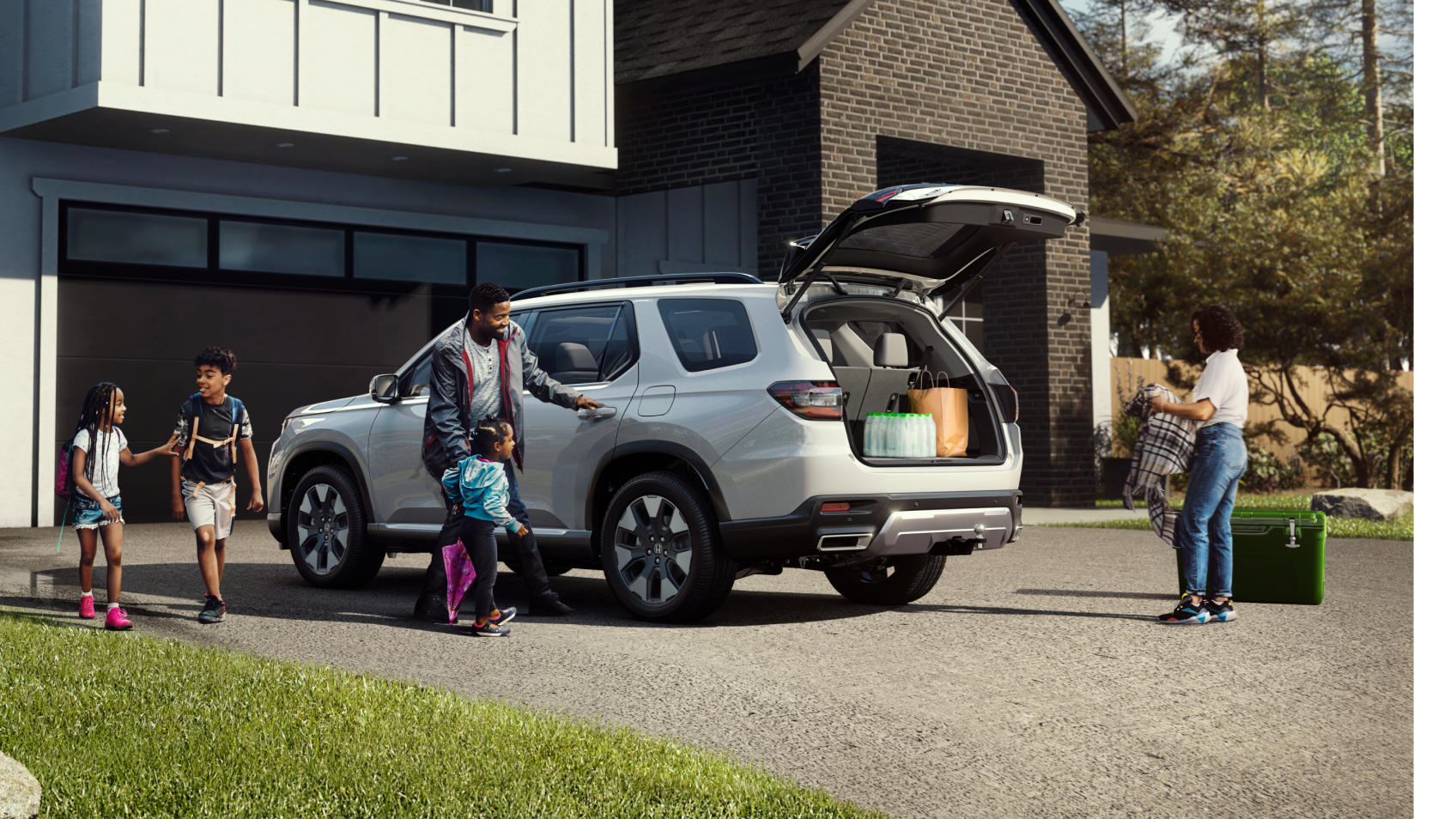 3/4 rear side view of a white Pilot parked in a driveway with its trunk open. Parents and three children are nearby, loading items and getting into the car.