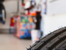 Closeup of a tire’s tread. In the background, out of focus, we see we’re in a mechanic’s shop.