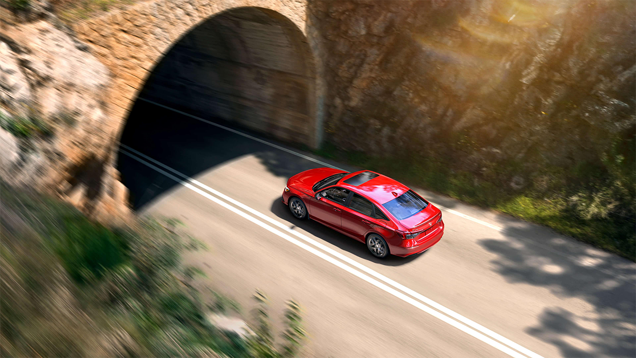 3/4 rear bird’s eye view of red Civic Sedan driving into a mountainside tunnel.