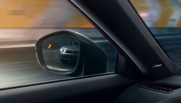 Interior closeup of the door mirror with the reflection of a car in it and a lit up blind spot indicator light.