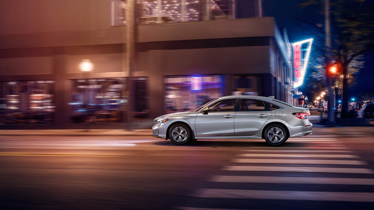Side wide view of grey Civic Sedan driving on a city street at night.