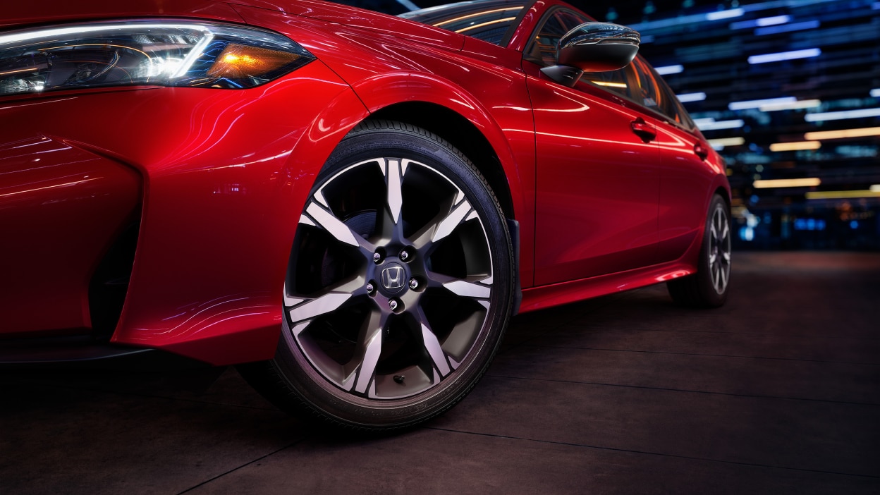 Closeup of the front wheel on a parked red Civic Sedan in a dark warehouse-like space.
