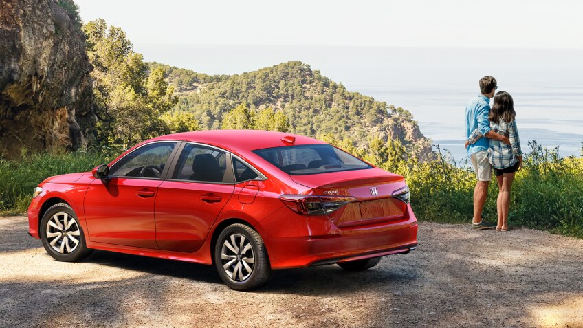 3/4 side rear view of red Civic Sedan parked at a scenic lookout spot overlooking plush green hills. A couple standing near it take in the scenery. 