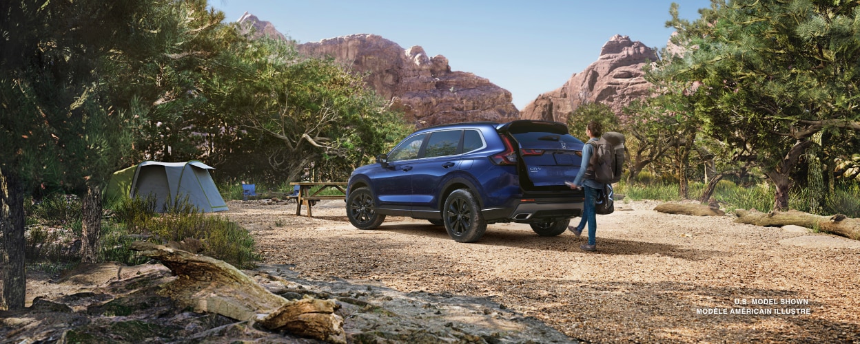 3/4 rear view of a blue CR-V parked in a desert campsite – woman with hiker’s backpack waving her foot under bumper sensor, opening the tailgate door.