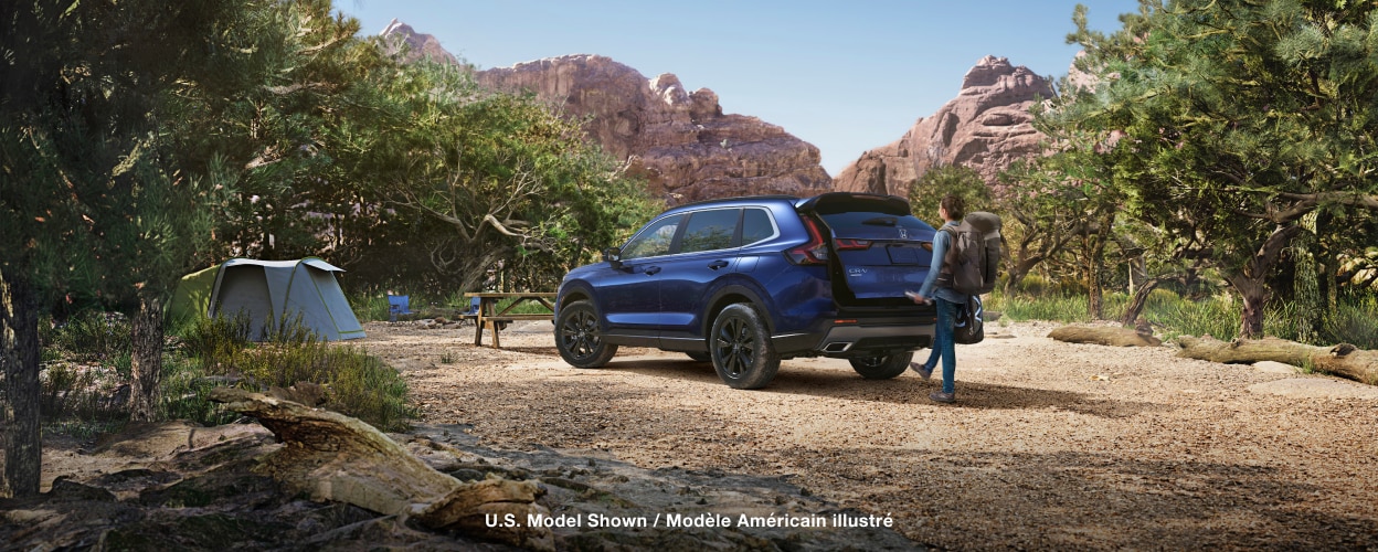 3/4 rear view of a blue CR-V parked in a desert campsite – woman with hiker’s backpack waving her foot under bumper sensor, opening the tailgate door.