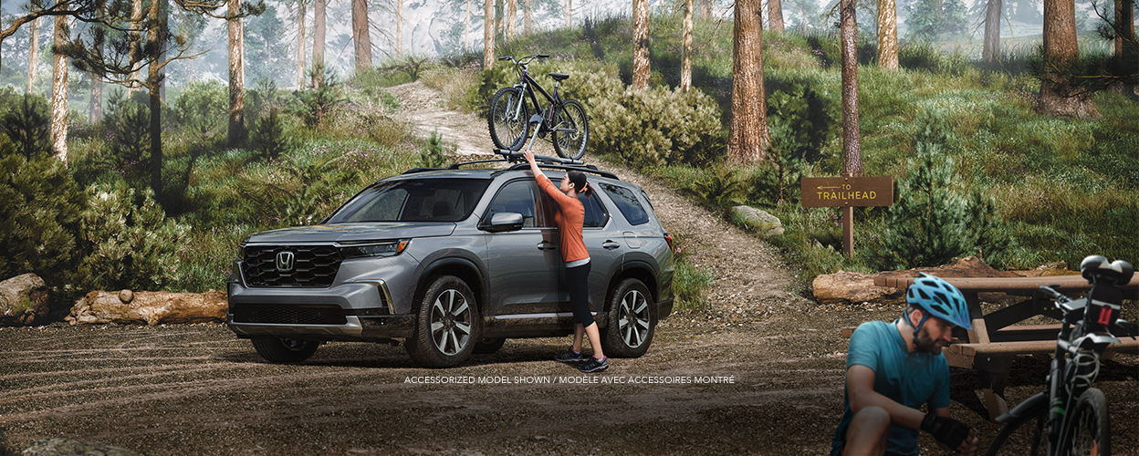 Wide sideview of grey Pilot, with a bike on the roof rack, towing a grey trailer uphill on a dirt road in a temperate rainforest.