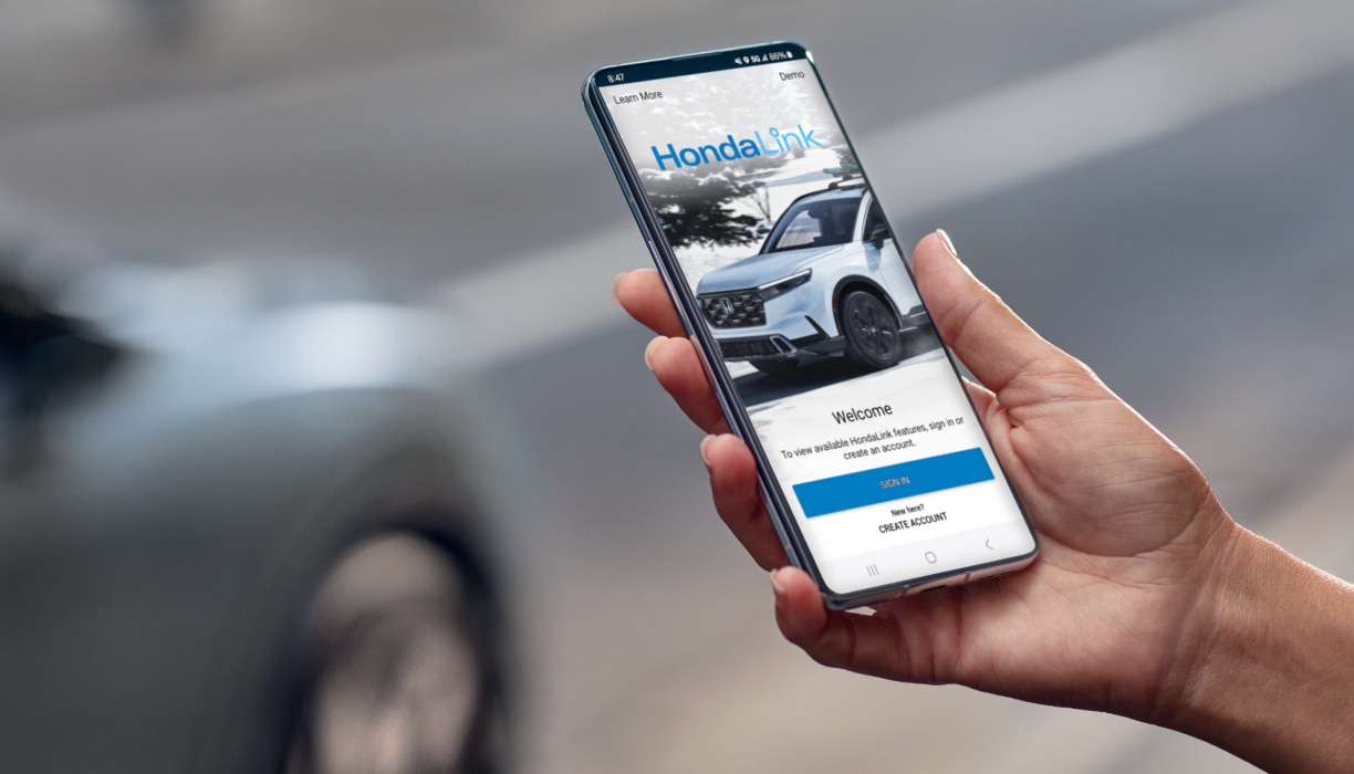 Closeup of hand using HondaLink phone app on a street. Parked grey vehicle in the background.