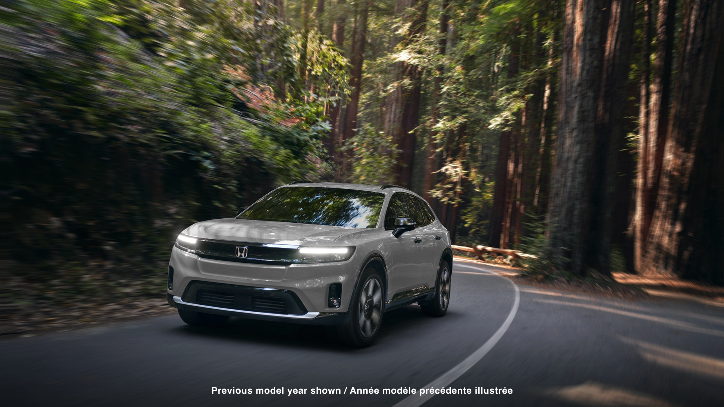 3/4 front view of white Prologue taking a turn on a paved road in a temperate forest.